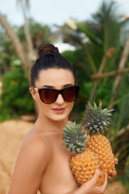 Güzel, neşeli, çıplak kız sahilde göğsünde ananas tutuyor. Bikinili bronz tenli kadın. Göğsünü ananasla kaplıyor.