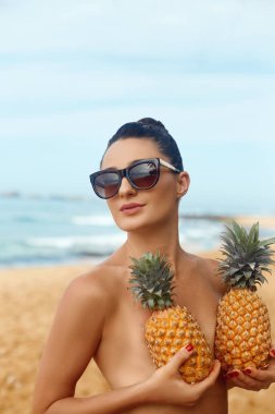 Yazın mavi gökyüzü ve okyanus arka planında koyu renkli mayosu olan güzel, beyaz tenli kadın model. Göğsünü ananasla kaplıyor.