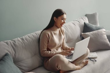 Dizüstü bilgisayar kullanan gülümseyen genç bir kadın, evdeki kanepede oturuyor, güzel bir kız alışveriş yapıyor ya da sosyal ağda sohbet ediyor, bilgisayar projesinde çalışan serbest çalışan biri.