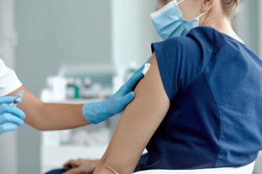 Tıbbi maske takan kadın hastanede iğne oluyor. Doktor ya da hemşire şırıngayı kadın hastaya iğne yapmaya hazırlıyor.