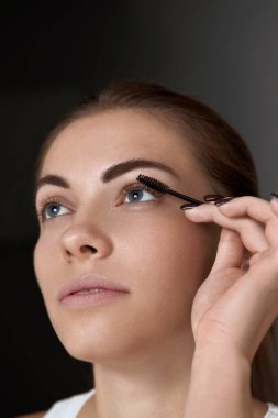 Mascara. Makyaj fırçasıyla kirpiklerine siyah rimel süren bir kadın. Güzel, genç bir kadın yüzü, doğal kaşları ve kirpikleriyle..  