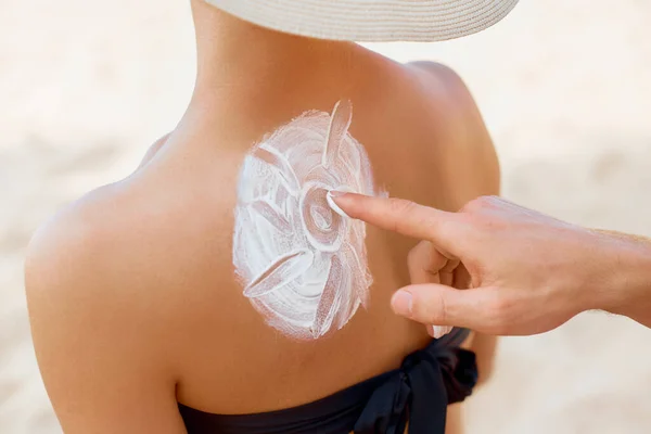 Man Applying Protective Sunscreen On Woman Tanned Shoulder In Form Of The Sun At Beach. Putting Sun Lotion On Female Skin. Skin And Body Care