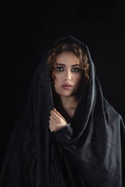 Koyu gri başörtülü güzel bir kadın yüzünü siyah arka planda lüks bir makyajla kapatıyor.
