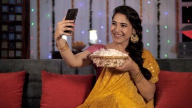 Çekici bir kadın, Hint Festivali 'nde cep telefonuyla selfie çekiyor. Bir sepet çiçekle kanepede oturan güzel bir kadın. Bayram sezonu, Diwali kutlamaları, ışık festivali, Hindu festivali.