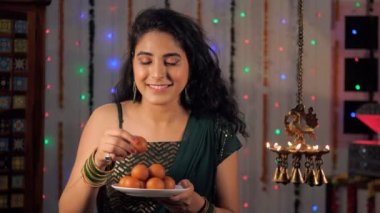 Genç bir kadın Diwali 'deki misafirlerine ev yapımı Gulaab Jamun ikram ediyor. Şenlik arka planında ışıl ışıl yanan güzel pirinç Diyas - bir Hindu festivali, hayırlı bir olay, olumlu titreşimler