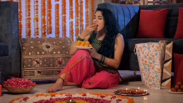 お祝いの季節に自家製ラドゥスを食べるのを楽しんでいる美しいインドの女の子 色とりどりの贈り物と燃えるダイヤが花の背景に一緒に保たれています Diwali祭り 縁起の良い機会 インド文化 — ストック動画