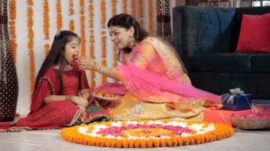 Gülümseyen genç anne, şenlikler sırasında tatlı kızına şeker yediriyor. Geleneksel Rangoli ve renkli bir hediye kutusu Diwali dekorasyonları, kutlama zamanı, bir Hindu festivali, aile ilişkileri.