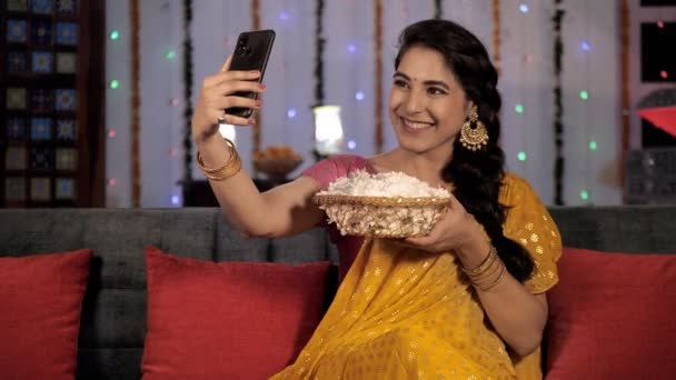 一个漂亮的女人在Sari用她的移动印度节点击一个自拍 漂亮的女性坐在沙发上 手里拿着一篮鲜花 节庆季节 Diwali庆典 印度教节日 — 图库视频影像