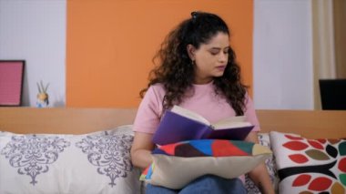 Yatak odasında otururken cep telefonuyla konuşan neşeli genç bir bayan. Uzun siyah saçlı, çekici bir ev kadınının evde kitap okuduğu yakın çekim - rahatlama ve rahatlama