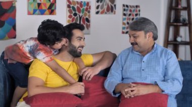 Günlük kıyafetler içinde, babası ve dedesiyle vakit geçiren genç bir çocuk. Aynı ailenin üç erkek üyesi bir kanepede oturuyor. Oturma odası, boş zaman, aile bağı, farklı nesiller.