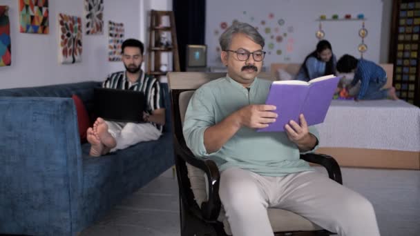 一位上了年纪的祖父 戴着一副眼镜 在家里看书 印度家庭 联合家庭 一位年轻的商人在笔记本电脑上工作 而他的妻子则在和他们可爱的儿子玩 遥远的工作 遥远的通信 — 图库视频影像
