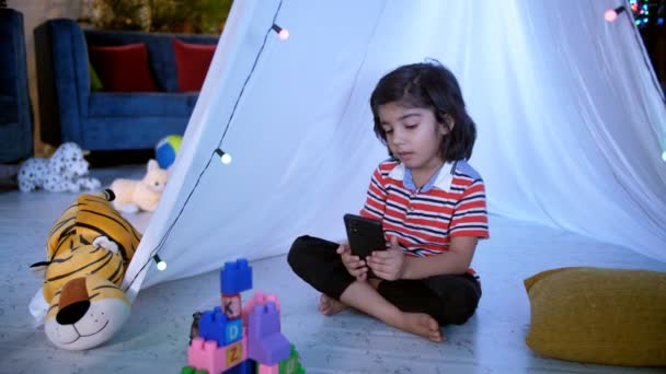 一个小男孩盘腿坐在地板上 在电话里看卡通片 一个穿着休闲装的小男孩坐在装饰着灯光的白色帐篷下 一个孩子 — 图库视频影像