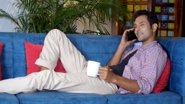 一个聪明的年轻人一边在手机上说话 一边在舒适的沙发上休息 一个穿着休闲装的帅哥 喝杯热茶 喝咖啡 闲暇时间 在电话里交谈 — 图库视频影像
