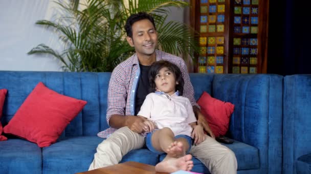 かわいい子供と彼のインドのお父さんは家で一緒にテレビを見ています 明るい父は彼がリビングルームで彼の膝の上に座っている間 彼の小さな息子をくすぐる 家族の絆 余暇時間 幸せな子供時代 — ストック動画