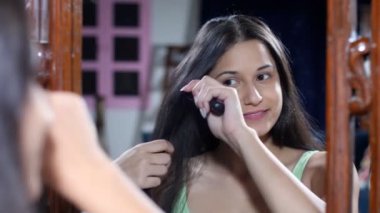 Siyah uzun saçlarını aynanın önünde tarayan, tarayan, gülümseyen genç bir kız. Güçlü ve sağlıklı saçlar, saç bakımı, saç maskesi, parlak saçlar. Neşeli bir bayan dışarı çıkmadan önce hazırlanıyor - moda, yaşam tarzı, bakım