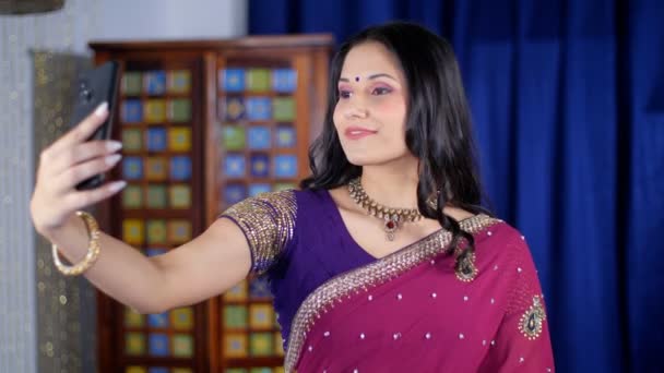 インドの文化 インドの結婚式 パーティーなど スマートフォンを使って自撮りをしながら楽しそうにポーズをとる魅力的な女性 インドの主婦は 彼女のリビングルームに立ってかなり見られ 宝石を身に着けています 民族服 手作りのジュエリー 現代的なライフ — ストック動画