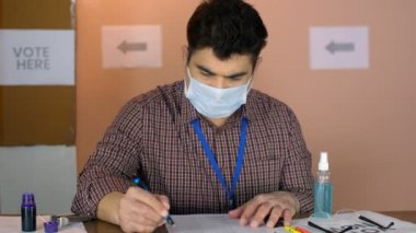 Tıbbi / yüz maskesi takan bir erkek subay seçim listelerini kontrol ediyor - Covid 19 yaşam tarzı, Coronavirus salgını, Corona sırasındaki anket. Yerel oy kullanma kabininde çalışan orta yaşlı bir Chunaav Adhikari - belediye seçimleri, oy sandığı, oy kullanma merkezi