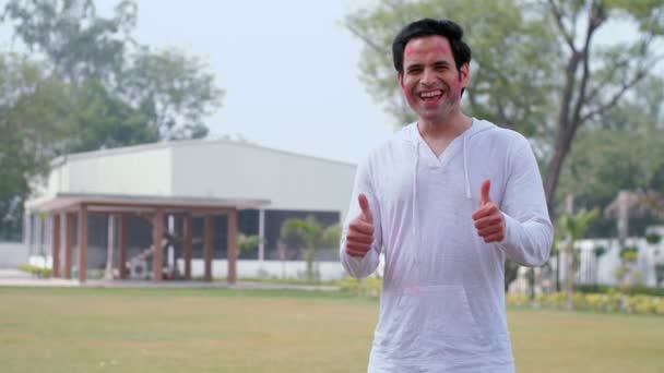 顔を有機的なガラルに覆われた若い男が親指アップジェスチャーをしています ホリ祭り 応援アップ 春祭り 鮮やかな色 公園でインドの色の祭りを祝う伝統的な服のハンサムな男 Festi — ストック動画