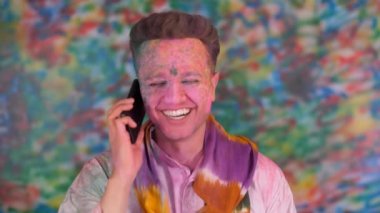 Holi 'yi kutlarken telefonda konuşan yakışıklı bir çocuk. Holi renklerine boyanmış bir yüz. Telefonda gülümseyen çekici genç yetişkin - Holi selamlar, renk festivali, renkli yüz