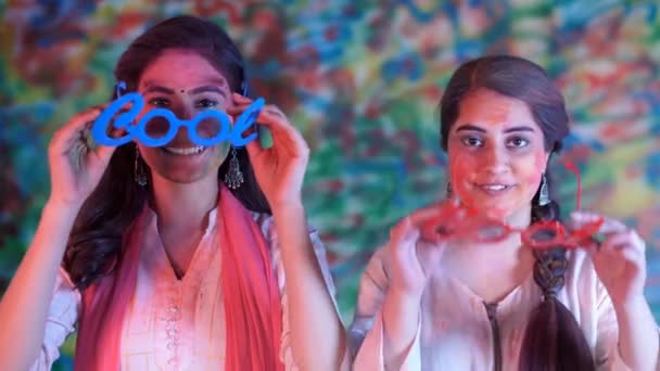 2人の笑顔のインドの女の子がカラフルな面白いゴーグルを着て ホリ祭りのカメラのポーズをとっています 美しい女性たちの顔は色で覆われ 両腕を組んで立っている — ストック動画