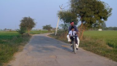 Kurta-pijama giyen mutlu bir çiftçi oğlunu bisiklete bindirip okula bırakıyor - köy eğitimi. Okuldan dönerken Hintli bir köyün üniformalı küçük bir çocuğu - Hindistan 'da okul eğitimi, köy eğitimi