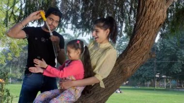 Çekici bir çift ve kızları sabun köpüğü üfleyip yakalıyorlar. Eğlenceli bir aktivite, çocukluk oyunu. Modern Hintli bir aile halka açık bir parkta vakit geçirir. Boş zaman, aile bağı, ilişki ve birliktelik.