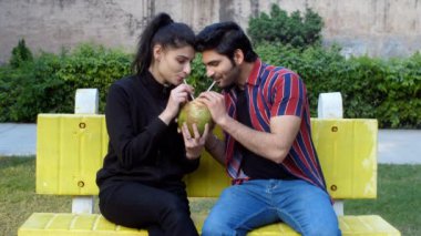 A cheerful Indian husband-wife drinking / sharing tender coconut water - a refreshing drink, a nutritious diet. A young romantic couple spending time in a park - relationship and togetherness