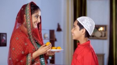 A pretty Muslim lady and her adorable son celebrate the festival of Eid together - an auspicious occasion, a festive season. A young mother feeds homemade Laddu to her little kid - family bonding, Indian sweets, a celebration time