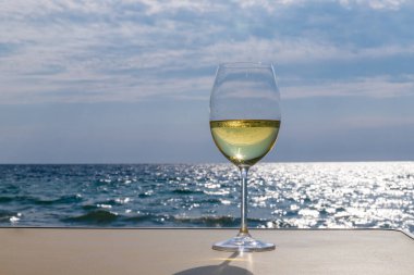 Bir bardak beyaz şarap. Deniz kıyısının arka planında şarap ve güneşli bir yaz plajı. Tatil köyünde şarap tatma ve rahatlama.