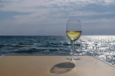 Bir bardak beyaz şarap. Deniz kıyısının arka planında şarap ve güneşli bir yaz plajı. Tatil köyünde şarap tatma ve rahatlama.