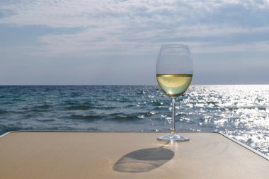Bir bardak beyaz şarap. Deniz kıyısının arka planında şarap ve güneşli bir yaz plajı. Tatil köyünde şarap tatma ve rahatlama.