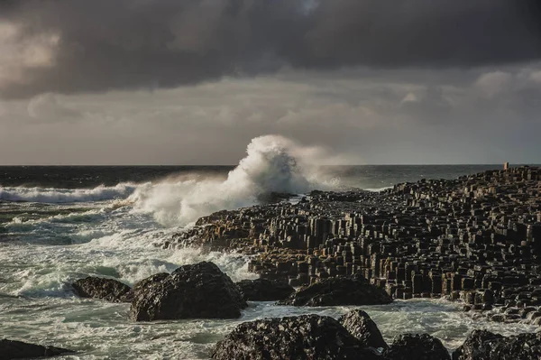 Rough ocean waves smashing on hexagonal rocks at the giant \'s causeway on a murky day. Hexagon - shaped stones with a sea wave hitting the cliff convey a strong impact or major, big effect concept. County Antrim, Northern Ireland, United Kingdom
