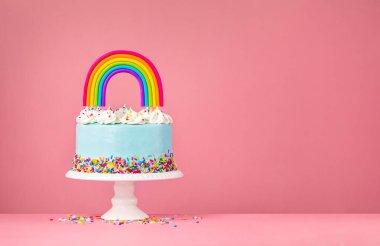 Mavi doğum günü pastası, krema süslemeleri, serpiştirmeler ve pembe arka planda renkli ev yapımı gökkuşağı süsü..