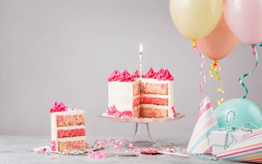 Dilimlenmiş pembe funfetti doğum günü pastası mum, hediyeler, şapkalar ve açık gri arka planda renkli balonlar. Bir doğum günü partisinden sahne!