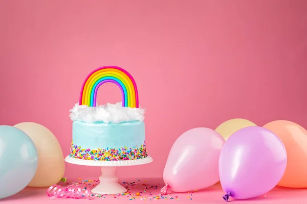 Torta di compleanno blu con arcobaleno e palloncini da festa su rosa Foto Stock