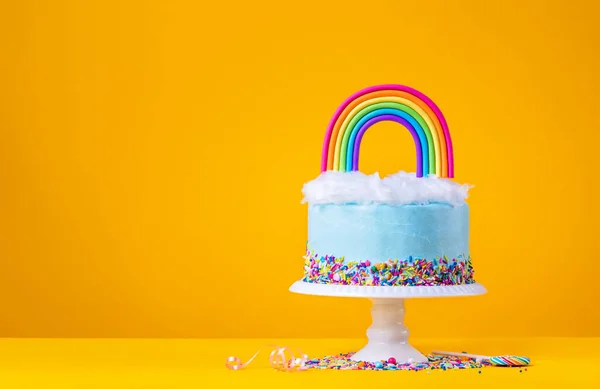 Blauwe verjaardagstaart met Rainbow topper op geel — Stockfoto