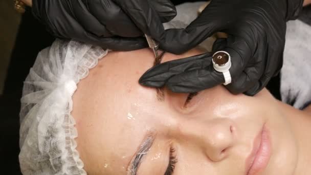 Kosmetikerin in schwarzen Handschuhen macht permanente Make-up-Korrektur der Form der Augenbrauen zu jungen schönen Mädchen. Microblading, Augenbrauen-Tätowierung im Schönheitssalon — Stockvideo