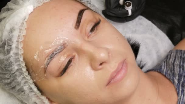 Kosmetolog i sorte handsker gør permanent makeup korrektion af øjenbrynenes form til ung smuk pige. Mikroblading, øjenbryn tatovering i en skønhedssalon – Stock-video