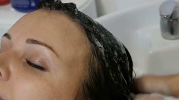 Mycie włosów w salonie. Specjalny umywalka i prysznic do mycia głowy. Młoda piękna kobieta z niebieskimi oczami i czarnymi włosami myć włosy u fryzjera — Wideo stockowe