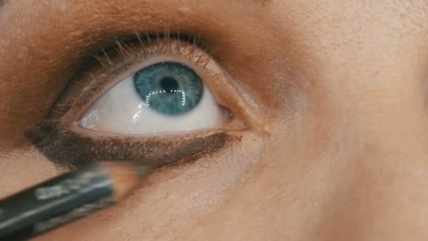 Επαγγελματική makeup artist κάνει μπεζ μακιγιάζ με ένα ειδικό μολύβι για μια νεαρή γυναίκα μοντέλο με μπλε μάτια. Βραδινό φωτεινό smoky μακιγιάζ ματιών — Αρχείο Βίντεο