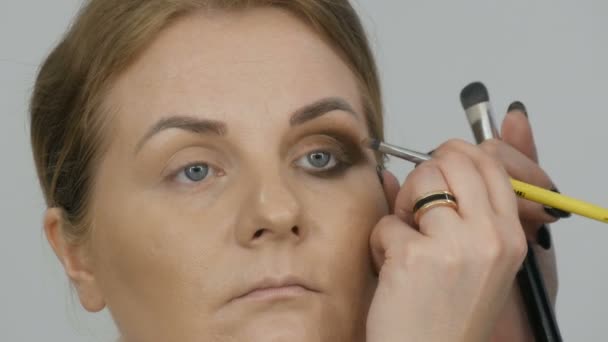 Επαγγελματίας μακιγιέρ κάνει μπεζ μακιγιάζ με ένα ειδικό πινέλο για μια νεαρή γυναίκα μοντέλο με μπλε μάτια. Βραδινό φωτεινό smoky μακιγιάζ ματιών — Αρχείο Βίντεο