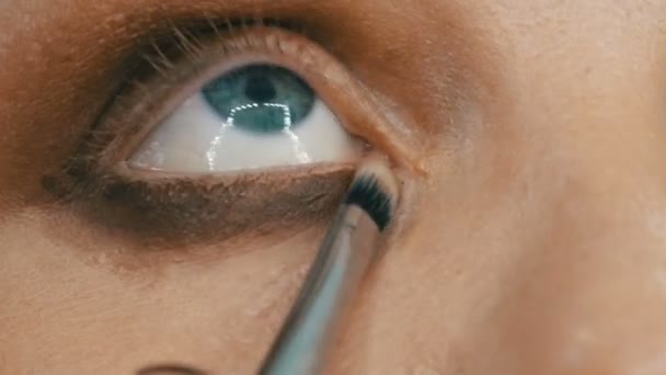 Επαγγελματίας μακιγιέρ κάνει μπεζ μακιγιάζ με ένα ειδικό πινέλο για μια νεαρή γυναίκα μοντέλο με μπλε μάτια από κοντά άποψη. — Αρχείο Βίντεο