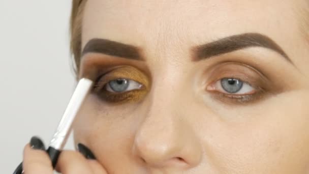 Master make-up artist tillämpa professionell make-up gyllene rökiga ögon i en skönhetssalong på en modell kvinna modell med blå ögon på nära håll — Stockvideo