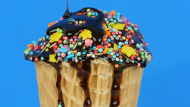 El glaseado de la salsa de chocolate y los coloridos espolvoreos fluyen sobre el helado en una taza de gofres sobre fondo azul — Vídeo de stock