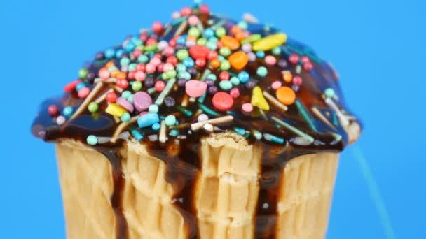 Cobertura de molho de chocolate e manchas coloridas flui sobre sorvete em uma xícara de waffle no fundo azul — Vídeo de Stock