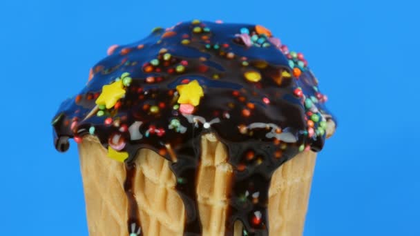 Schokolade Sauce Zuckerguss und bunte Streusel fließt über Eis in einem Waffelbecher auf blauem Hintergrund — Stockvideo