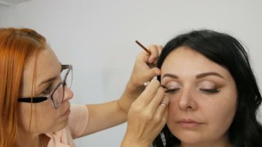 Bej renkli göz makyajı ile günlük moda. Özel fırçaların yardımıyla stilisti güzel mavi gözlü genç bir kadın modeline göz makyajı ve kaş şekli düzeltmesi yapıyor.