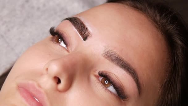 Master anvender hvide markeringer til korrekt form af øjenbryn med en speciel børste. Det smukke ansigt af ung pige model på sofaen i en skønhedssalon før microblading, permanent makeup øjenbryn tatovering – Stock-video