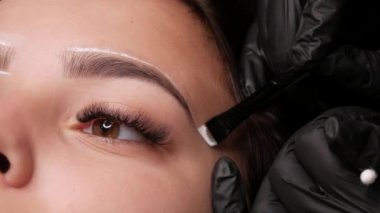 Usta beyaz işaretleri özel bir fırçayla kaşların şeklini düzeltmek için uygular. Mikroplu patinaj yapmadan önce güzellik salonundaki kanepede oturan genç bir kızın güzel yüzü, kalıcı makyaj kaşlı dövme.