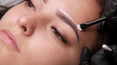 Usta beyaz işaretleri özel bir fırçayla kaşların şeklini düzeltmek için uygular. Mikroplu patinaj yapmadan önce güzellik salonundaki kanepede oturan genç bir kızın güzel yüzü, kalıcı makyaj kaşlı dövme.